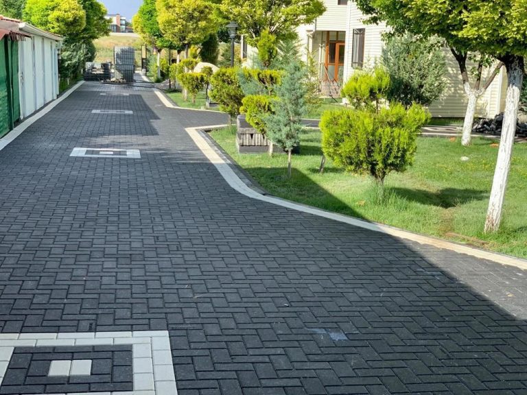 Koyu renkli kilitli parke taşları ve beyaz detaylarla döşenmiş, ağaçlı bir bahçe yolu.