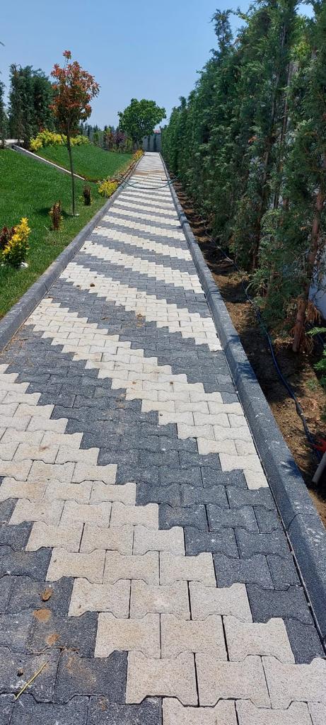 Kırmızımsı kilitli parke taşlarıyla döşenmiş, ağaçlarla çevrili kıvrımlı bir patika yolu.