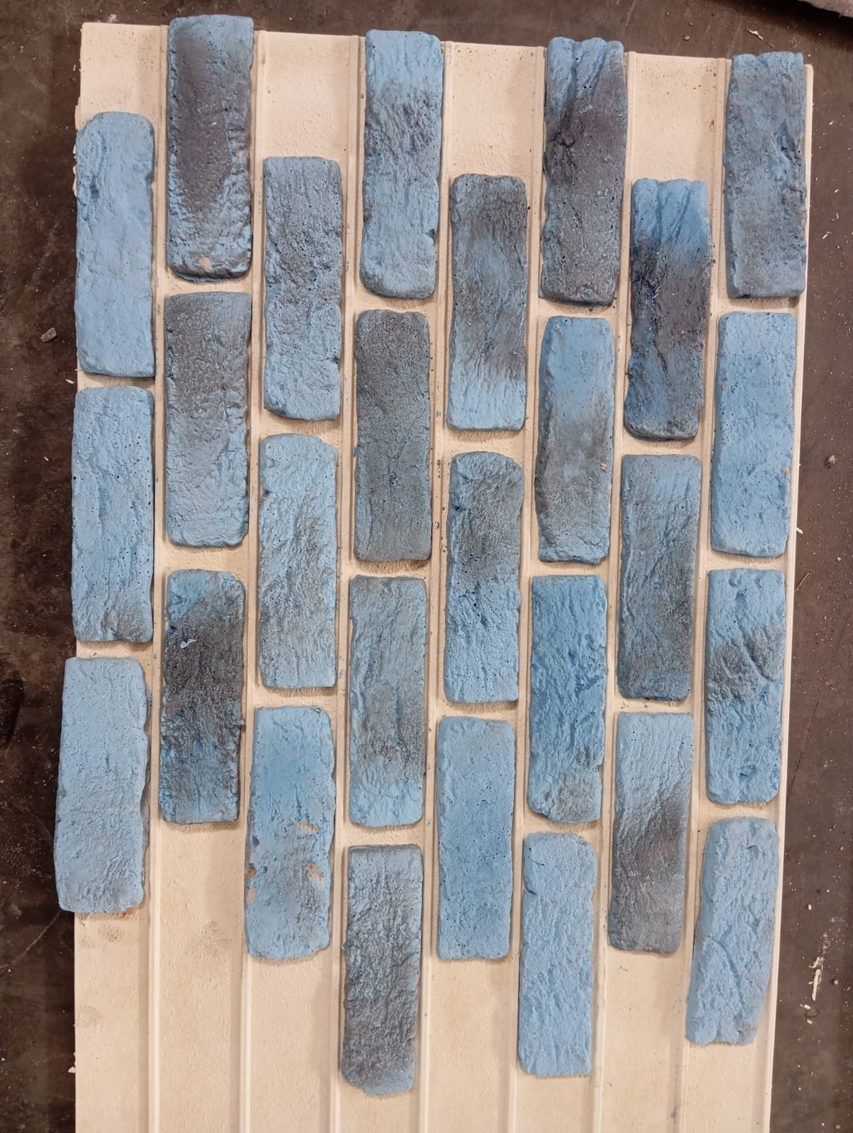 Açık renkli bir panel üzerinde dikey dizilmiş, mavi ve gri tonlarında eskitme yüzeyli dekoratif kültür tuğlaları.